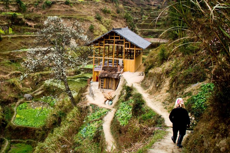 A little girl peering at her grandmother walking towards a rustic home in Miao village.