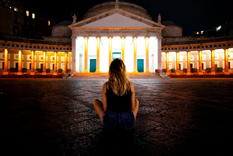 A figure seated in front of illuminated historic architecture in Naples at night.