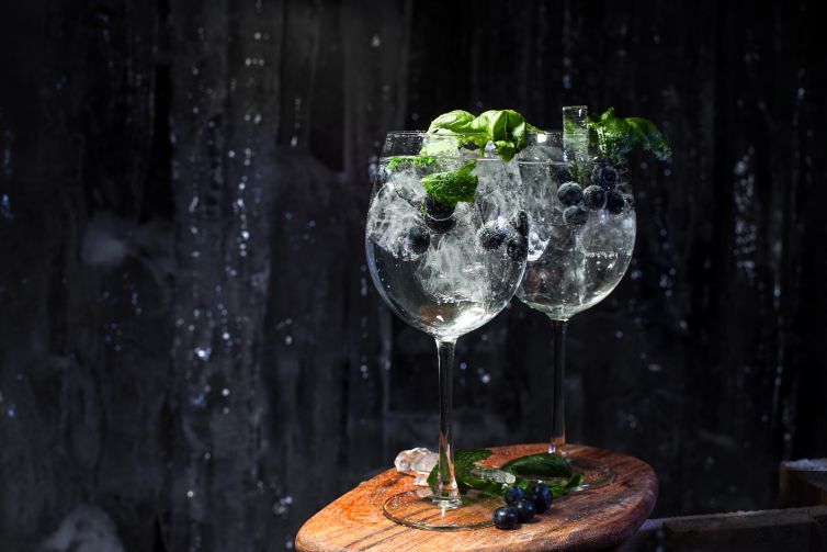 Two glasses of mint and blueberry drinks on a wooden surface with ice.