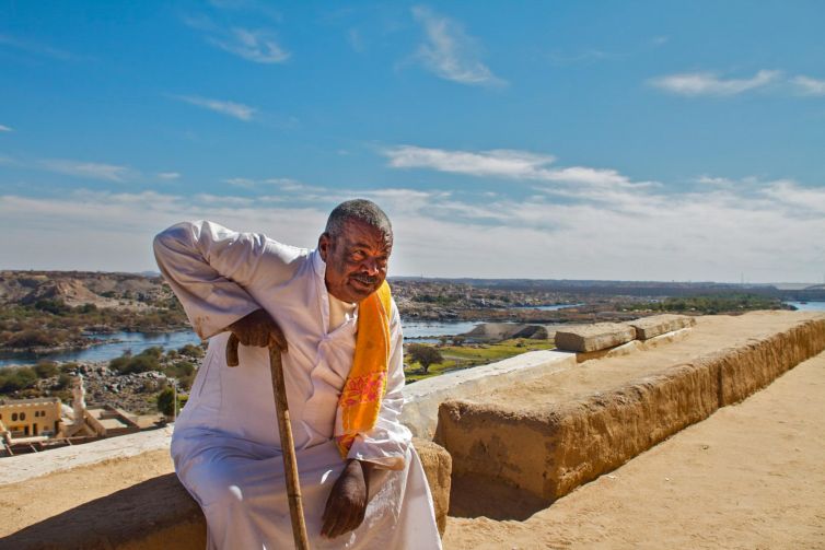 Man in traditional attire with cane overlooking the Nile River and Aswan landscape.