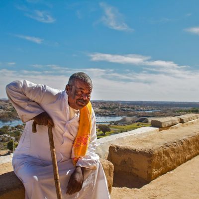 Man in traditional attire with cane overlooking the Nile River and Aswan landscape.
