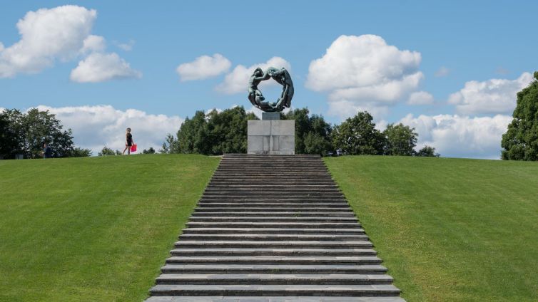Sculpture on a hill in Oslo with people walking nearby and clouds in the sky.