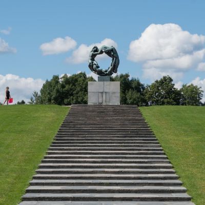 Sculpture on a hill in Oslo with people walking nearby and clouds in the sky.